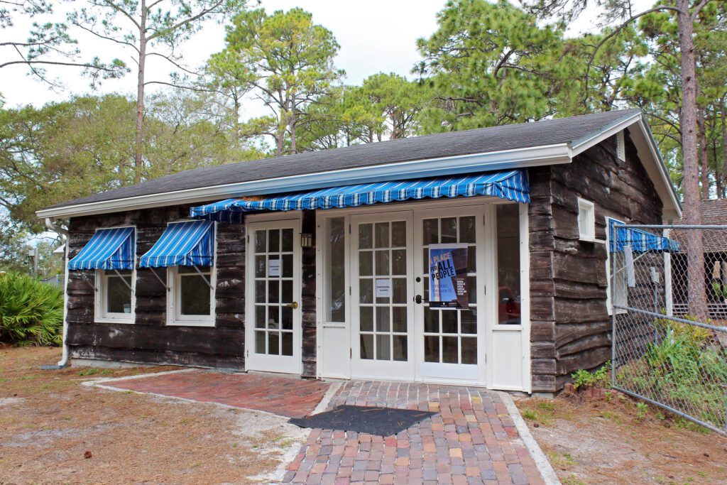 A quaint wooden building with blue and white striped awnings over the windows and a matching entrance canopy, featuring a white door with a sign, surrounded by greenery under a cloudy sky.