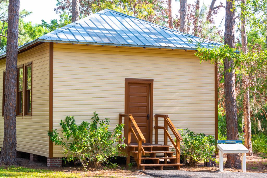 A small yellow building with a metal roof, wooden stairs, surrounded by trees and greenery on a sunny day, with an information signpost to the side.