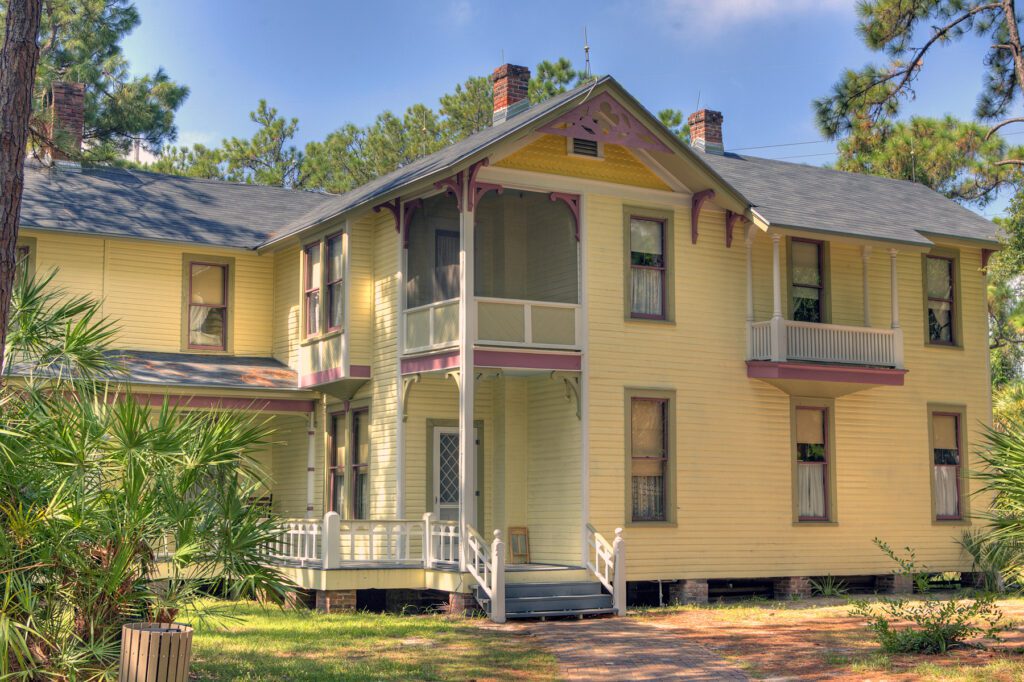 A charming yellow two-story house with a wraparound porch nestled among pine trees on a sunny day.